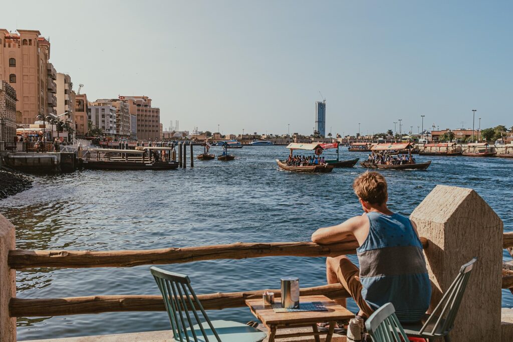 dubai creek, waterfront, man sitting, abra boats, nature, traditional boats, dubai water taxis, relaxing view, outdoor seating, creek view, people, abra ride, sunny day, sightseeing, waterfront cafe, boat ride, casual scene, cityscape, urban view, waterfront dining, riverfront, dubai, pier, sea, outdoors, boat
